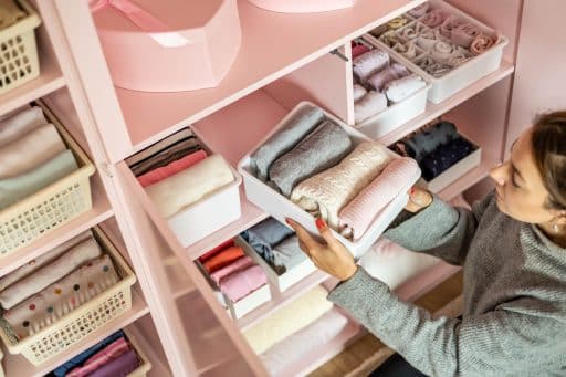 Mulher organizando roupas em um armário rosa, utilizando caixas organizadoras para dobrar e armazenar peças de forma eficiente. O ambiente é bem iluminado e apresenta uma disposição harmoniosa dos itens.