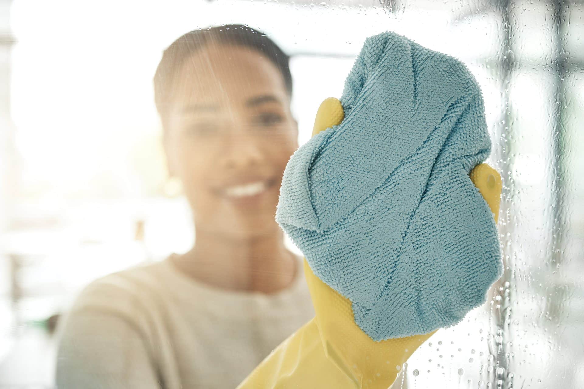 Mulher sorridente usando luvas de borracha amarelas limpa um vidro com um pano de microfibra azul. O vidro tem gotas de água, indicando um processo de higienização. O fundo está desfocado, sugerindo um ambiente iluminado e arejado.