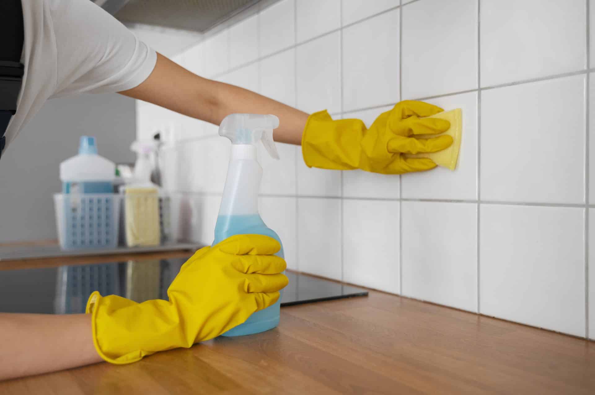 A imagem mostra uma pessoa limpando azulejos brancos em uma cozinha. Ela usa luvas de borracha amarelas para proteção e segura um borrifador com líquido azul em uma das mãos, enquanto esfrega os azulejos com uma esponja na outra. Sobre a bancada de madeira, é possível ver outros produtos de limpeza organizados em um cesto ao fundo.