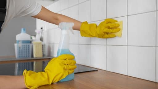 A imagem mostra uma pessoa limpando azulejos brancos em uma cozinha. Ela usa luvas de borracha amarelas para proteção e segura um borrifador com líquido azul em uma das mãos, enquanto esfrega os azulejos com uma esponja na outra. Sobre a bancada de madeira, é possível ver outros produtos de limpeza organizados em um cesto ao fundo.