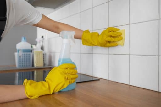 A imagem mostra uma pessoa limpando azulejos brancos em uma cozinha. Ela usa luvas de borracha amarelas para proteção e segura um borrifador com líquido azul em uma das mãos, enquanto esfrega os azulejos com uma esponja na outra. Sobre a bancada de madeira, é possível ver outros produtos de limpeza organizados em um cesto ao fundo.