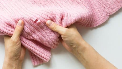 O ChatGPT disse: A imagem mostra um par de mãos segurando uma peça de roupa de tricô rosa, que apresenta um buraco visível causado por traças. A malha do tecido está danificada, com fios soltos ao redor da área afetada, indicando o desgaste provocado por insetos. A cena destaca a fragilidade de tecidos armazenados inadequadamente e reforça a importância de cuidados preventivos para evitar danos por pragas.
