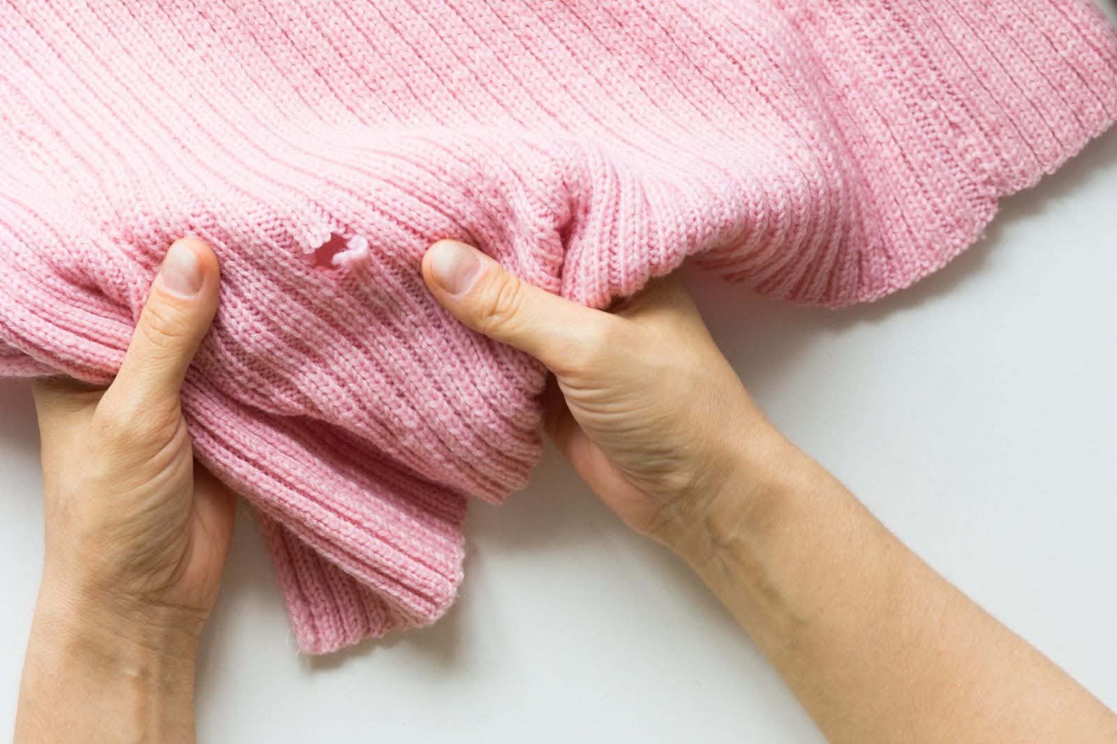 O ChatGPT disse: A imagem mostra um par de mãos segurando uma peça de roupa de tricô rosa, que apresenta um buraco visível causado por traças. A malha do tecido está danificada, com fios soltos ao redor da área afetada, indicando o desgaste provocado por insetos. A cena destaca a fragilidade de tecidos armazenados inadequadamente e reforça a importância de cuidados preventivos para evitar danos por pragas.