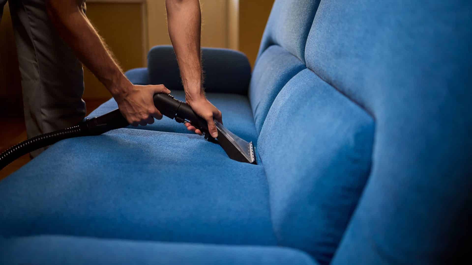 A imagem mostra uma pessoa realizando a limpeza profunda de um sofá azul com o auxílio de um equipamento extrator. O profissional utiliza um bico específico para estofados, aplicando sucção para remover sujeira e umidade dos tecidos. A ação é feita com precisão, focando nos vãos entre os assentos.