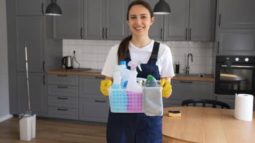 A imagem mostra uma diarista sorridente, vestindo uniforme azul e luvas de borracha amarelas, segurando uma cesta organizada com produtos de limpeza, como borrifadores, desinfetantes e panos. Ela está posicionada no centro de uma cozinha moderna com armários cinza, bancada de madeira clara e piso laminado. O ambiente transmite sensação de ordem, limpeza e profissionalismo nos serviços domésticos.