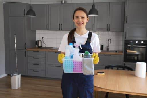 A imagem mostra uma diarista sorridente, vestindo uniforme azul e luvas de borracha amarelas, segurando uma cesta organizada com produtos de limpeza, como borrifadores, desinfetantes e panos. Ela está posicionada no centro de uma cozinha moderna com armários cinza, bancada de madeira clara e piso laminado. O ambiente transmite sensação de ordem, limpeza e profissionalismo nos serviços domésticos.