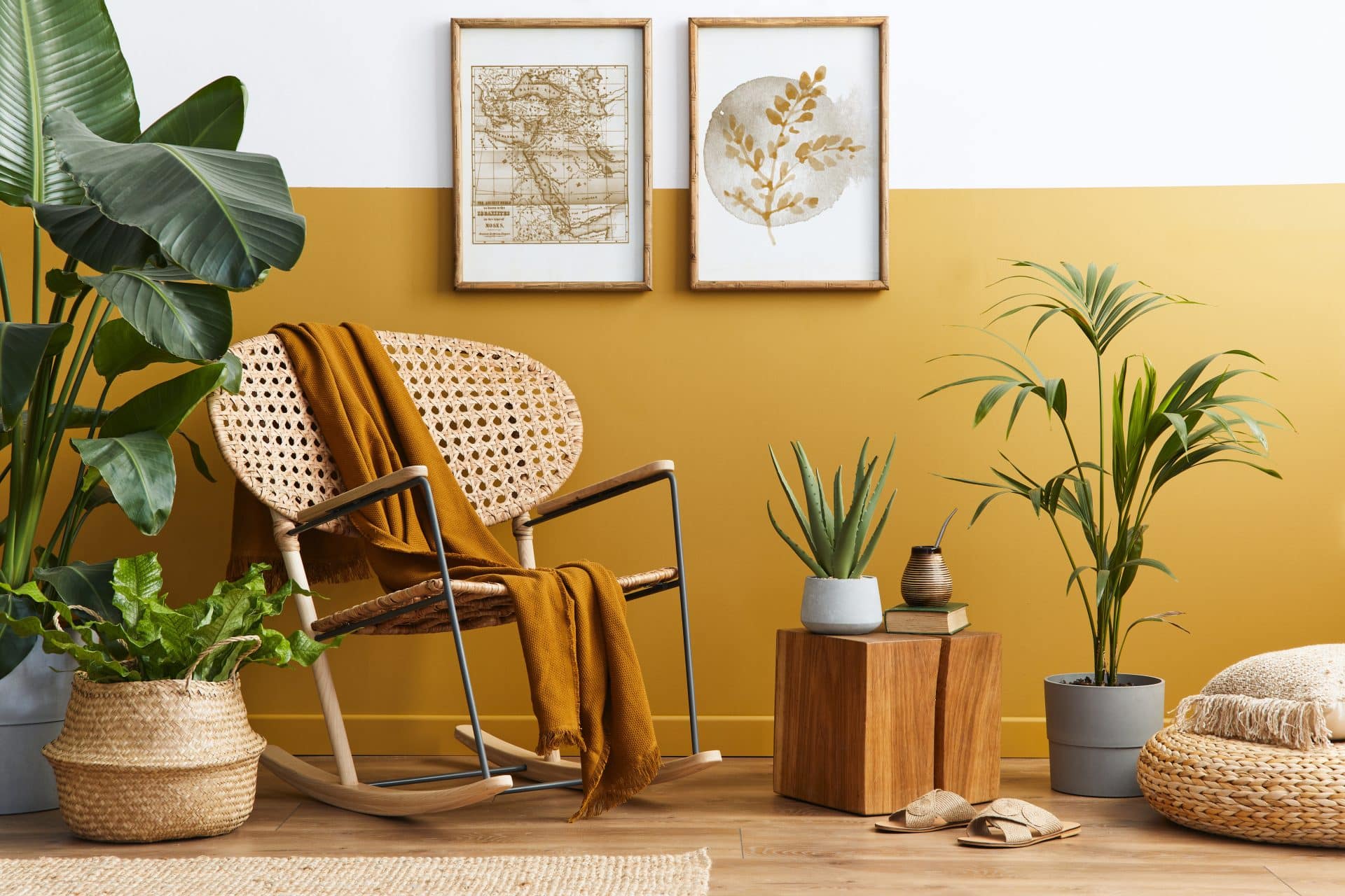 A imagem retrata um ambiente de decoração aconchegante e natural, com estilo boho-chic. Em destaque, uma cadeira de balanço em madeira e palha, sobre a qual repousa uma manta mostarda, harmonizando com a meia parede pintada em tom amarelo-ocre. Ao redor, diversas plantas em vasos de cerâmica e palha reforçam a atmosfera orgânica, junto a elementos decorativos como um pufe trançado, um banquinho de madeira, livros e uma cuia com bomba.