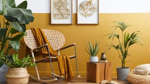 A imagem retrata um ambiente de decoração aconchegante e natural, com estilo boho-chic. Em destaque, uma cadeira de balanço em madeira e palha, sobre a qual repousa uma manta mostarda, harmonizando com a meia parede pintada em tom amarelo-ocre. Ao redor, diversas plantas em vasos de cerâmica e palha reforçam a atmosfera orgânica, junto a elementos decorativos como um pufe trançado, um banquinho de madeira, livros e uma cuia com bomba.