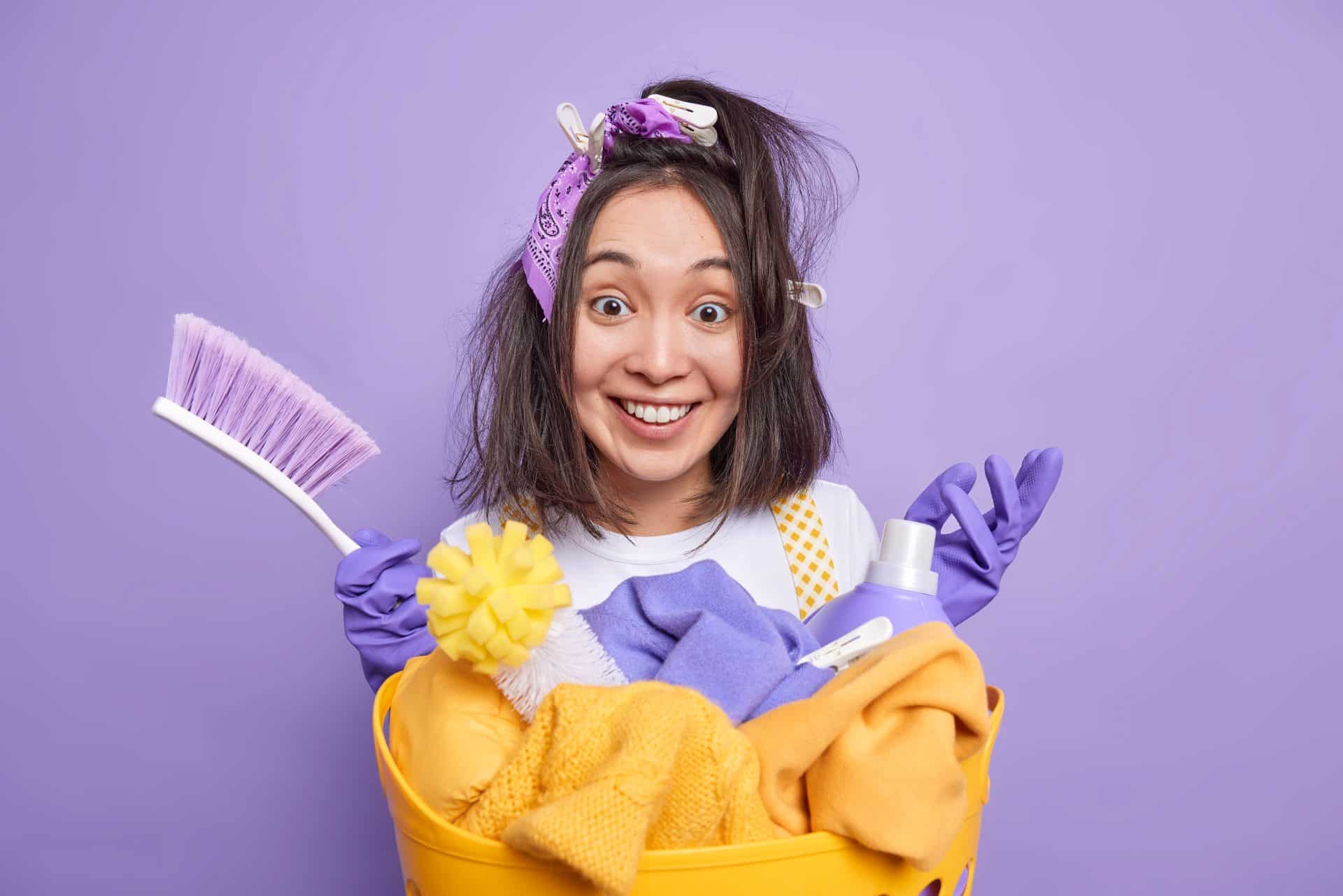 A imagem mostra uma mulher sorridente com aparência divertida e bagunçada, usando luvas de limpeza roxas, um lenço na cabeça e grampos de roupa no cabelo. Ela segura uma cesta amarela cheia de itens de limpeza, como panos, escova, esponja e um frasco de produto, todos em tons de amarelo e lilás. O fundo também é lilás, criando um visual vibrante e descontraído, que transmite o espírito leve e bem-humorado de uma rotina de faxina.