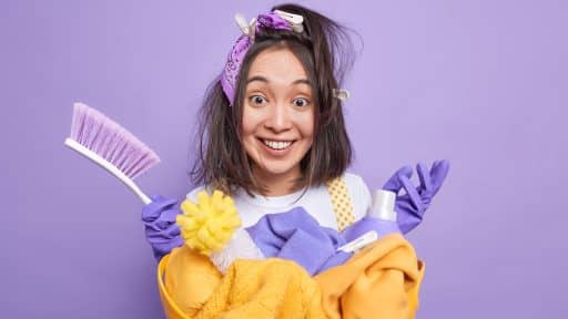 A imagem mostra uma mulher sorridente com aparência divertida e bagunçada, usando luvas de limpeza roxas, um lenço na cabeça e grampos de roupa no cabelo. Ela segura uma cesta amarela cheia de itens de limpeza, como panos, escova, esponja e um frasco de produto, todos em tons de amarelo e lilás. O fundo também é lilás, criando um visual vibrante e descontraído, que transmite o espírito leve e bem-humorado de uma rotina de faxina.
