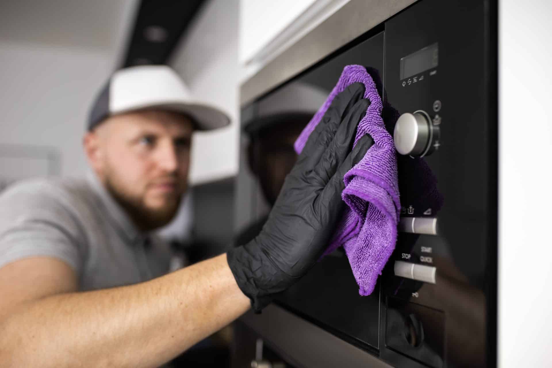 A imagem mostra um homem realizando a limpeza de um eletrodoméstico moderno, possivelmente um forno ou micro-ondas embutido. Ele usa uma luva preta de proteção e um pano de microfibra roxo para higienizar a superfície de vidro e aço inoxidável do aparelho, demonstrando cuidado com a manutenção e a preservação do equipamento. O ambiente é uma cozinha bem iluminada e organizada, sugerindo atenção aos detalhes e boas práticas de limpeza doméstica.