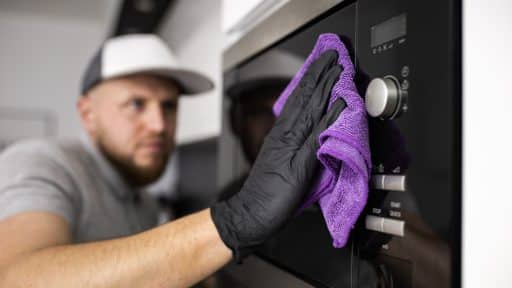 A imagem mostra um homem realizando a limpeza de um eletrodoméstico moderno, possivelmente um forno ou micro-ondas embutido. Ele usa uma luva preta de proteção e um pano de microfibra roxo para higienizar a superfície de vidro e aço inoxidável do aparelho, demonstrando cuidado com a manutenção e a preservação do equipamento. O ambiente é uma cozinha bem iluminada e organizada, sugerindo atenção aos detalhes e boas práticas de limpeza doméstica.