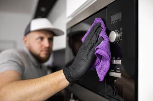 A imagem mostra um homem realizando a limpeza de um eletrodoméstico moderno, possivelmente um forno ou micro-ondas embutido. Ele usa uma luva preta de proteção e um pano de microfibra roxo para higienizar a superfície de vidro e aço inoxidável do aparelho, demonstrando cuidado com a manutenção e a preservação do equipamento. O ambiente é uma cozinha bem iluminada e organizada, sugerindo atenção aos detalhes e boas práticas de limpeza doméstica.