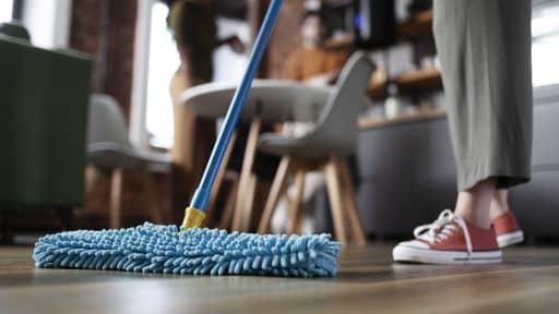 Pessoa limpando o chão com esfregão de microfibra azul em ambiente doméstico, com outras duas pessoas desfocadas ao fundo.