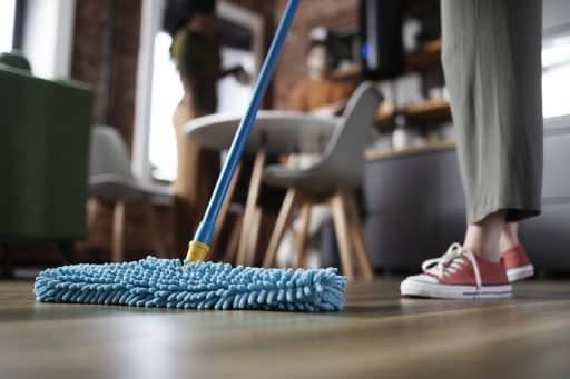 Pessoa limpando o chão com esfregão de microfibra azul em ambiente doméstico, com outras duas pessoas desfocadas ao fundo.