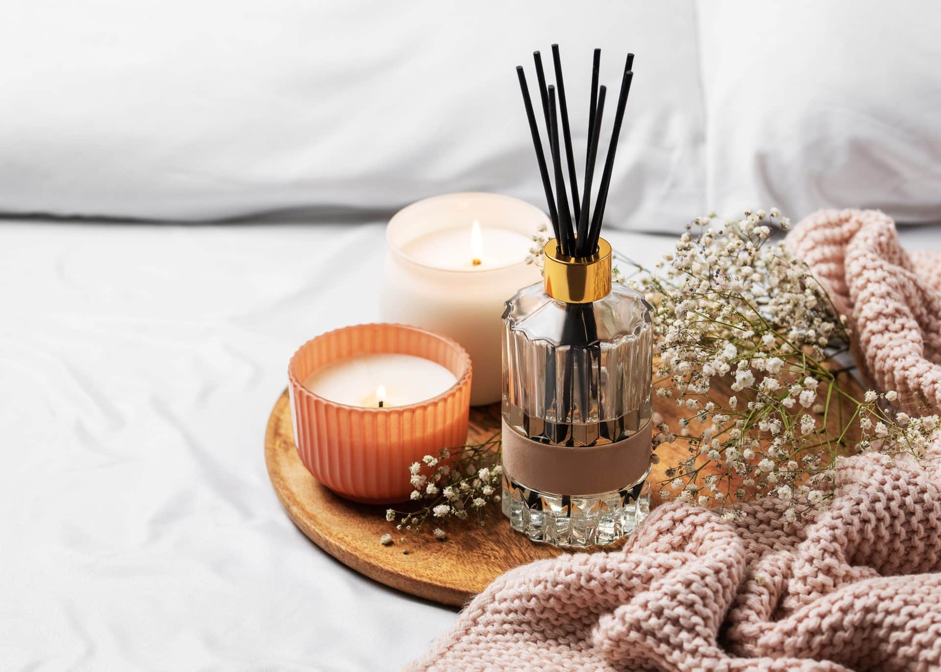 Bandeja de madeira com velas acesas, difusor de aromas, flores delicadas e manta rosa sobre uma cama, criando clima aconchegante e relaxante.