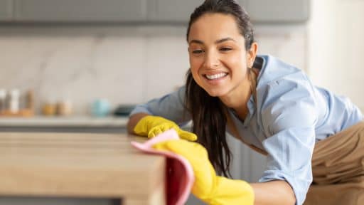 Mulher sorridente usando luvas amarelas e pano rosa, limpando uma superfície de madeira em cozinha.