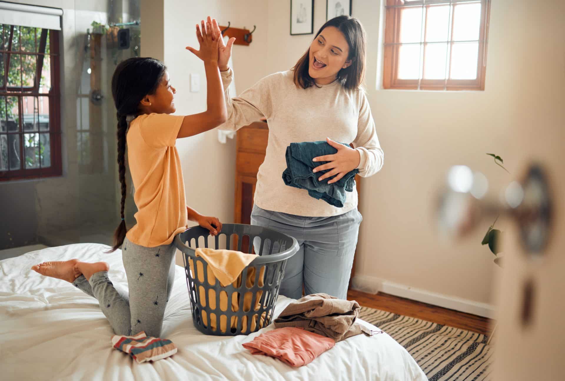Mãe e filha dando um "high five" enquanto dobram roupas juntas sobre a cama, com clima de cooperação e alegria.