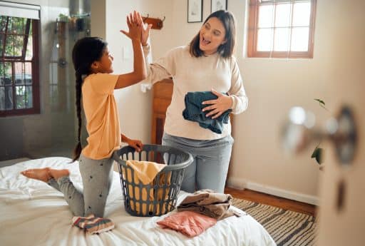 Mãe e filha dando um "high five" enquanto dobram roupas juntas sobre a cama, com clima de cooperação e alegria.