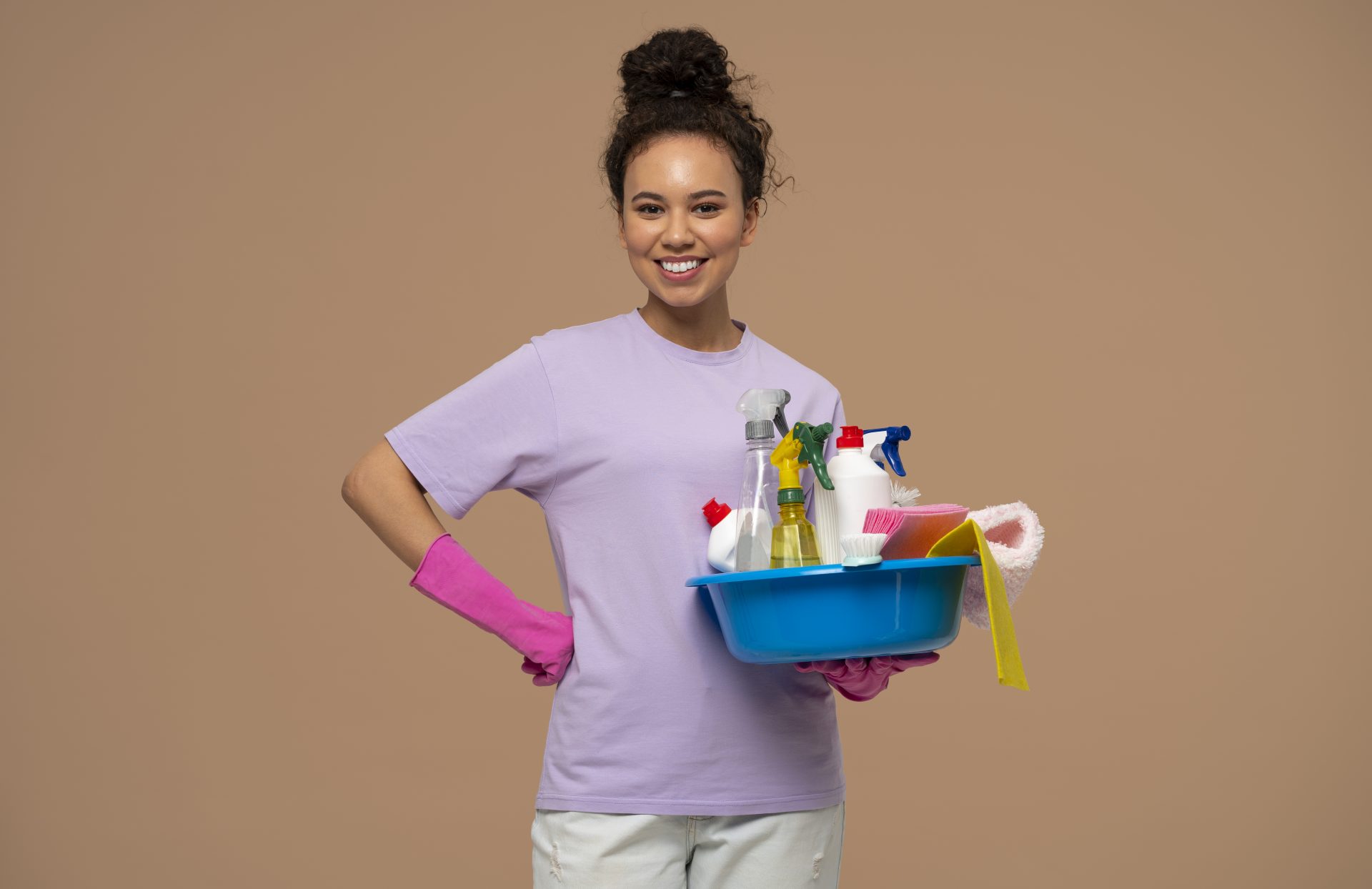 Mulher sorridente com luvas rosas segurando bacia azul cheia de produtos de limpeza, em fundo neutro.