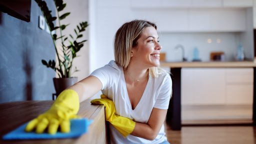 Mulher sorridente limpando móvel com pano azul e luvas amarelas em cozinha moderna.