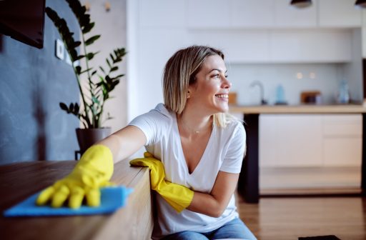 Mulher sorridente limpando móvel com pano azul e luvas amarelas em cozinha moderna.