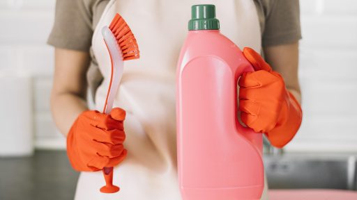 Pessoa com luvas laranja segurando escova de limpeza e frasco grande de detergente rosa.