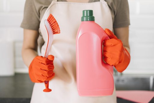 Pessoa com luvas laranja segurando escova de limpeza e frasco grande de detergente rosa.