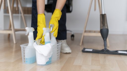 Pessoa com luvas amarelas pegando cesto com produtos de limpeza sobre piso de madeira clara.