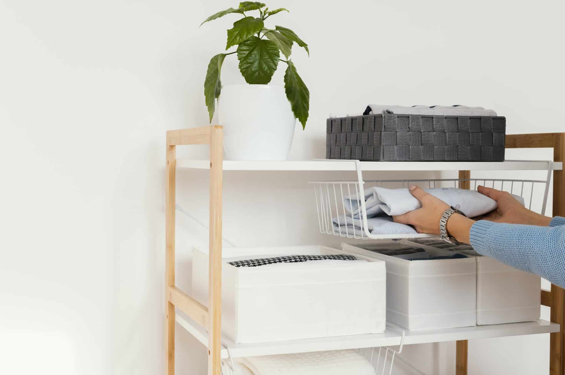 Pessoa organizando roupas dobradas em prateleira com cestos e caixas organizadoras, com vaso de planta decorativa no topo.