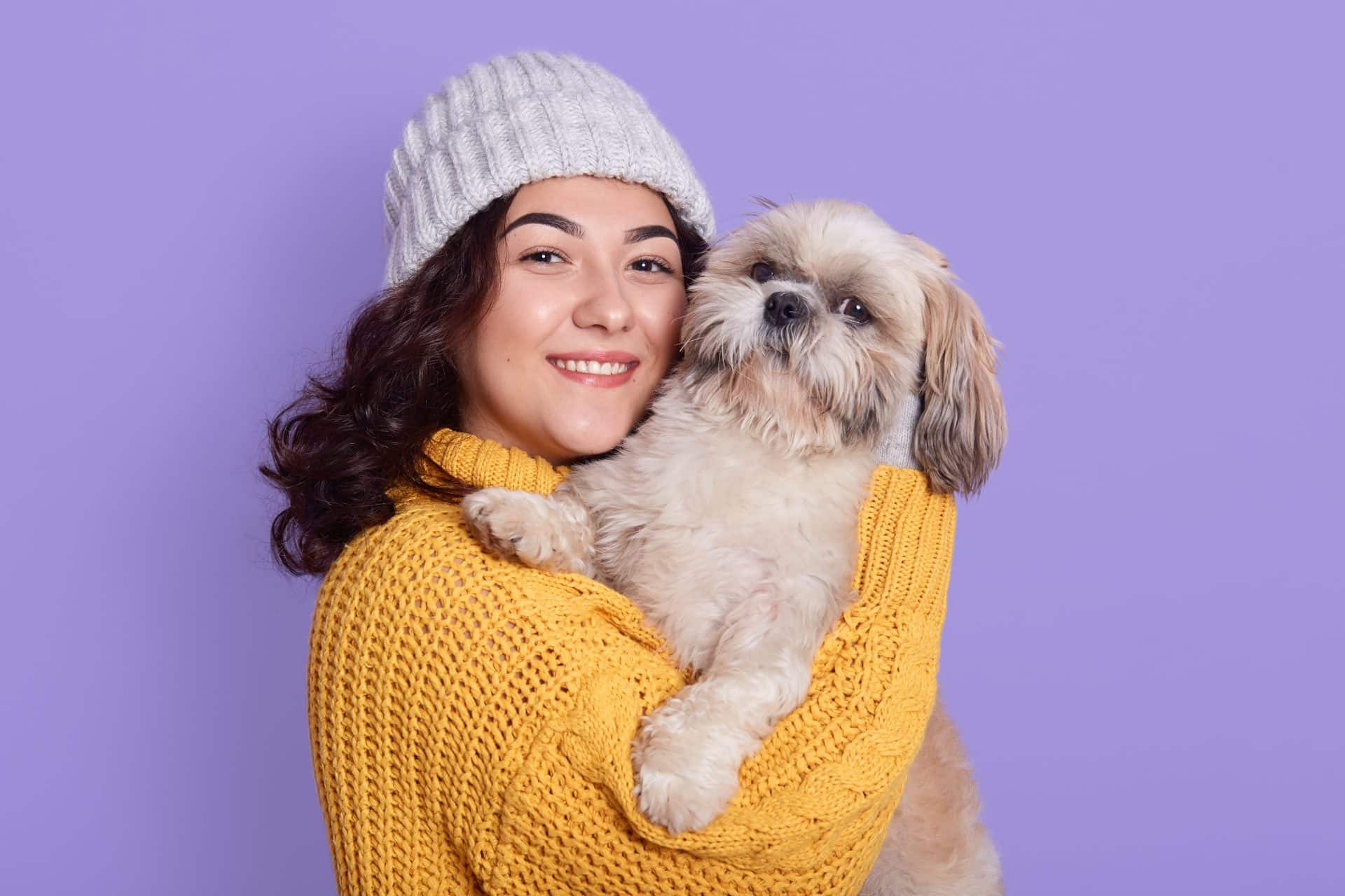 Mulher sorridente vestindo suéter amarelo e touca cinza segurando um cachorro da raça Shih-tzu em frente a fundo lilás.
