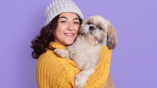 Mulher sorridente vestindo suéter amarelo e touca cinza segurando um cachorro da raça Shih-tzu em frente a fundo lilás.