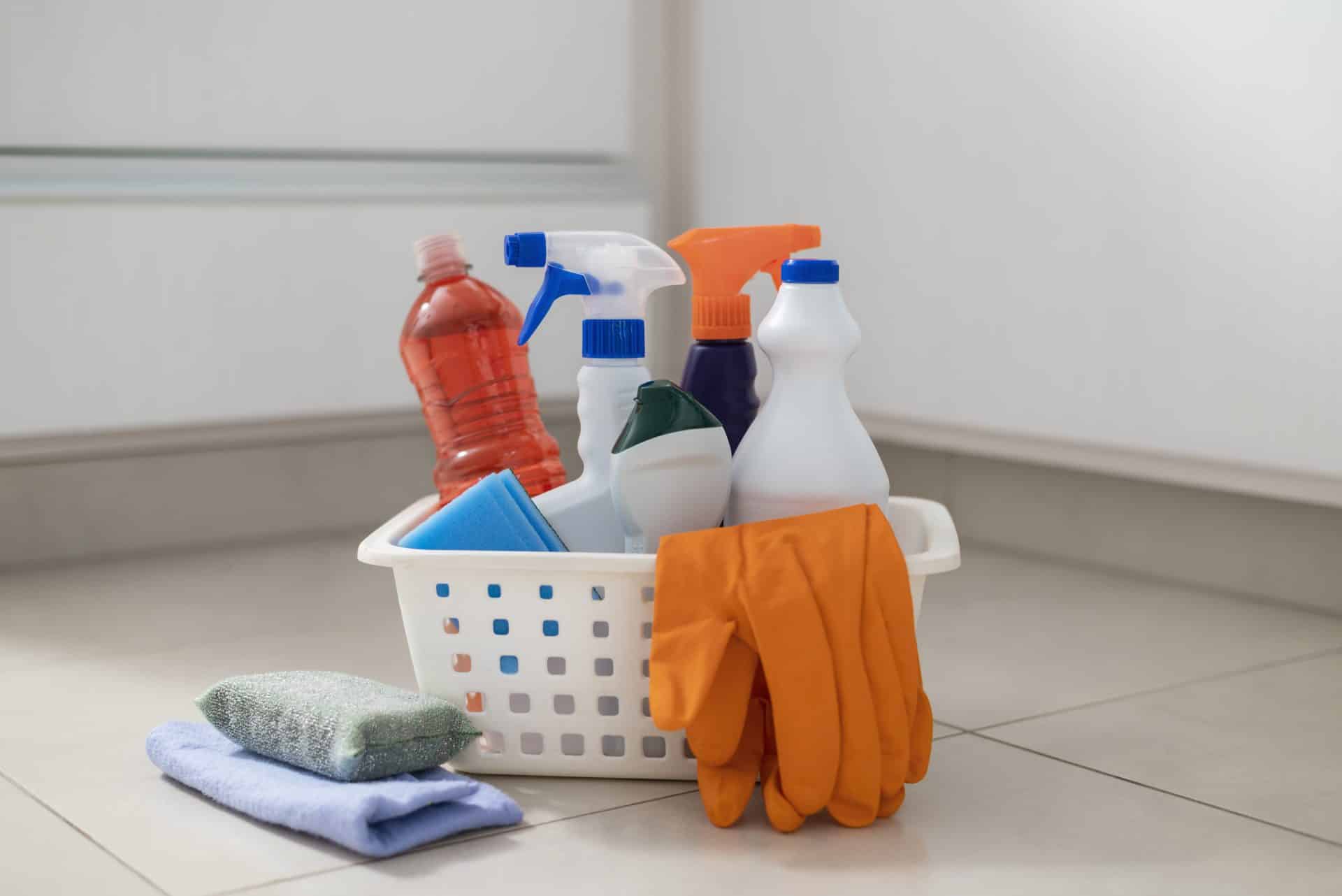 Cesto branco com produtos de limpeza variados, panos, esponja e par de luvas de borracha laranja, sobre piso claro.