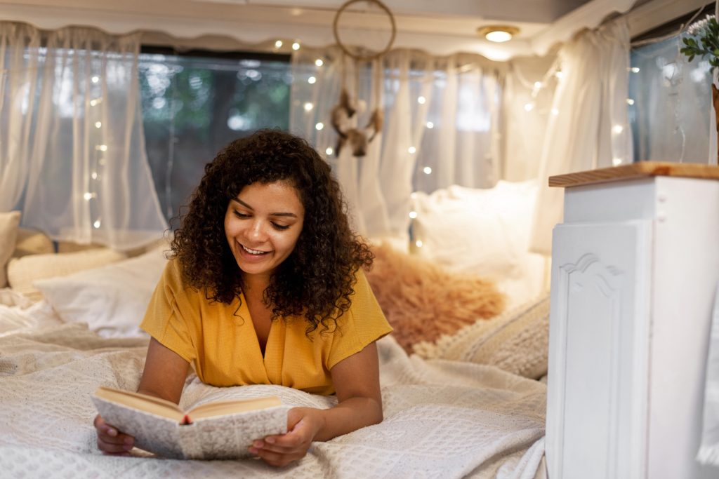 Mulher deitada em cama aconchegante, lendo um livro, com luzes decorativas ao fundo e atmosfera acolhedora.