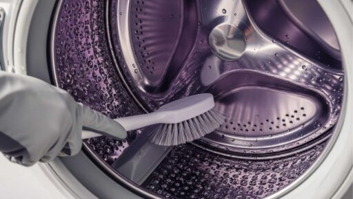 Pessoa com luva cinza limpa o interior do tambor de uma máquina de lavar com escova.