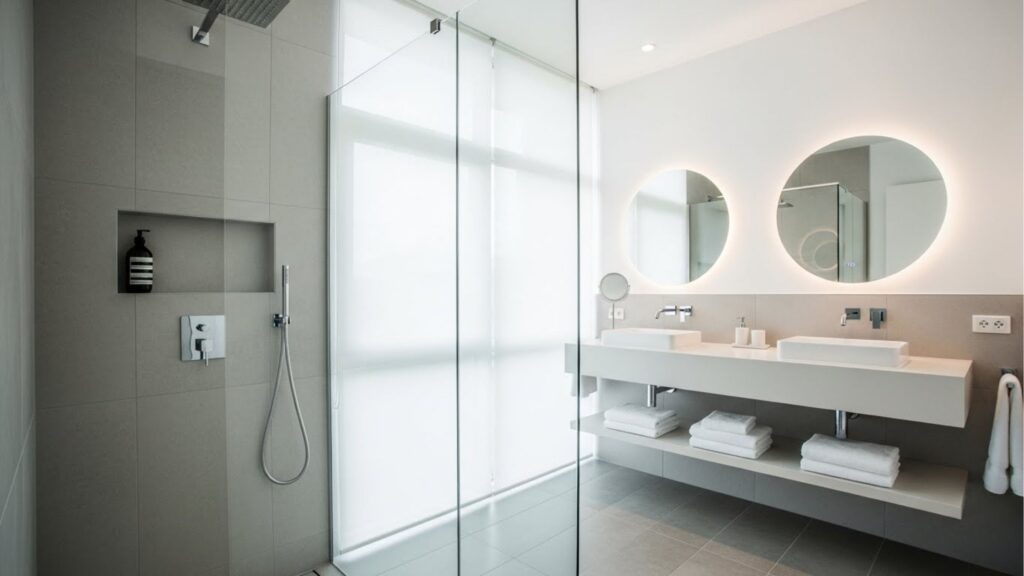 Banheiro moderno com box de vidro, chuveiro embutido no teto e nicho com sabonete líquido; ao fundo, bancada com duas cubas, espelhos redondos iluminados e toalhas dobradas.