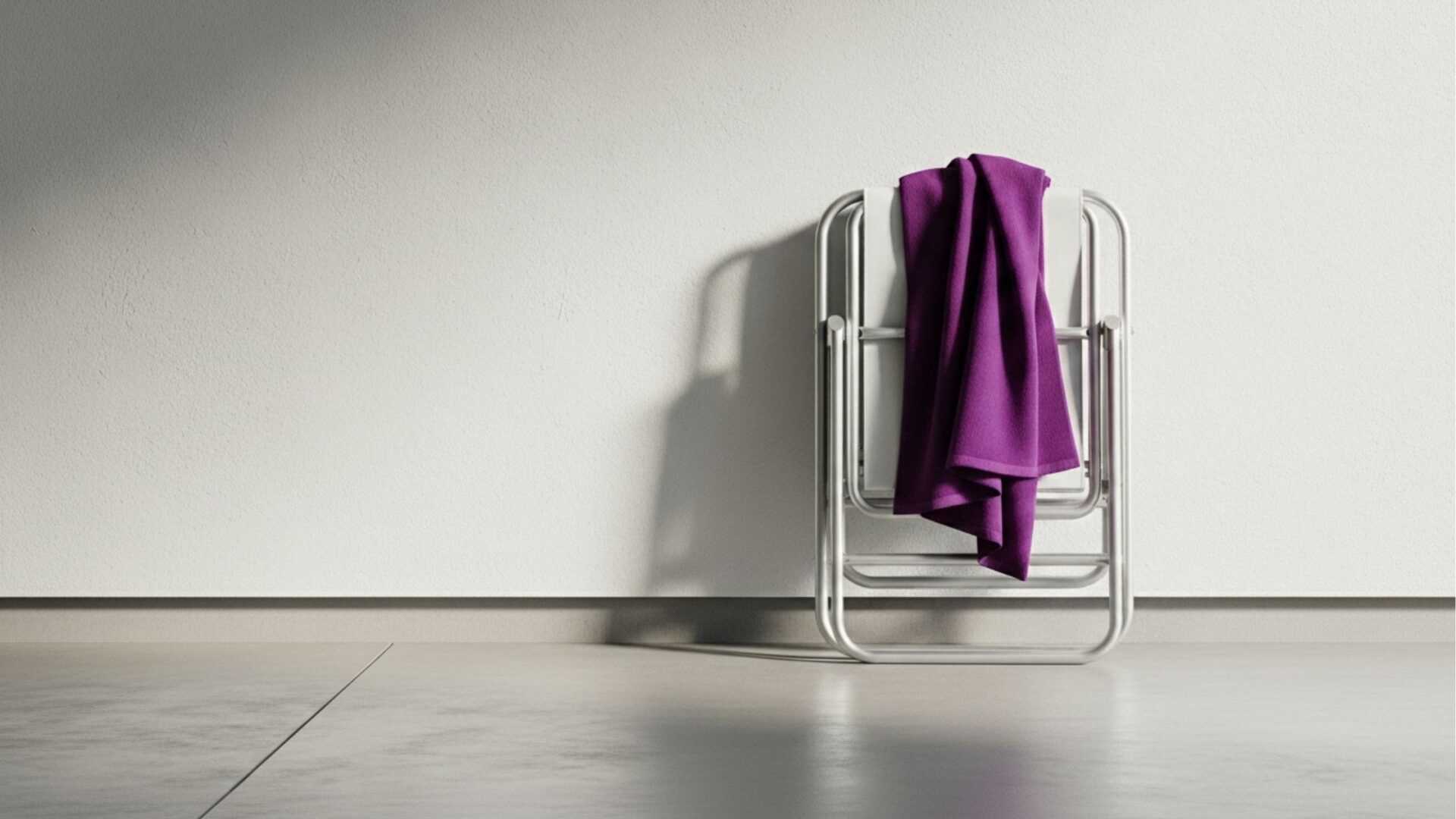 Cadeira de praia de alumínio dobrada e encostada em uma parede branca em ambiente interno, com uma toalha roxa pendurada nela.