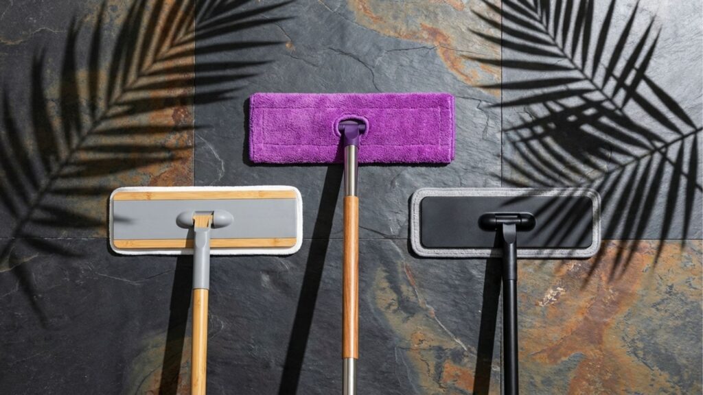 Uma visão superior plana de três esfregões de chão modernos organizados lado a lado em um piso de pedra texturizada escura com veios enferrujados. Os esfregões estão em formação de V invertido. O esfregão da esquerda tem um cabo de madeira clara, uma cabeça cinza com detalhes de bambu e uma almofada branca. O esfregão central tem um cabo de metal e madeira com uma almofada de microfibra roxa texturizada e brilhante. O esfregão da direita tem um cabo e uma cabeça pretos foscos com uma almofada preta. Sombras dramáticas de folhas de palmeira tropicais são projetadas no piso de pedra, enquadrando os esfregões.