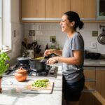 Mulher sorridente cozinhando em uma cozinha iluminada, com um vaso de manjericão fresco no balcão, uma das plantas que espantam insetos naturalmente em ambientes internos.