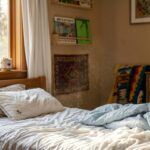 Uma foto de um quarto infantil aconchegante e desarrumado. Um forte feixe de luz solar dourada entra pela janela de madeira à esquerda, revelando inúmeras partículas de poeira suspensas no ar acima de uma cama com lençóis brancos e edredom azul bagunçado. Livros e brinquedos de madeira, incluindo um urso de pelúcia, estão visíveis nas prateleiras e na cômoda ao fundo.