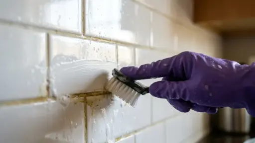 Um close-up extremo de uma mão usando uma luva de borracha roxa esfregando rejunte sujo e amarelado entre azulejos de metrô brancos molhados com uma pequena escova de limpeza preta. A superfície dos azulejos está coberta por espuma de limpeza branca e água, evidenciando a sujeira acumulada sendo removida. A ação de esfregar cria mais espuma ao redor da escova e do rejunte. O fundo está completamente desfocado, mantendo o foco nítido na mão, na escova e nos azulejos em processo de limpeza. (limpeza profunda em casa)