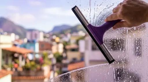Close-up de uma mão usando um rodo de metal com cabo roxo para limpar uma janela de vidro coberta de água com sabão. O movimento do rodo revela uma faixa do vidro perfeitamente limpa e transparente. Ao fundo, uma paisagem urbana ensolarada com montanhas aparece bastante desfocada. (como limpar vidros perfeitamente)