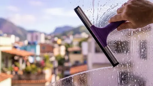 Close-up de uma mão usando um rodo de metal com cabo roxo para limpar uma janela de vidro coberta de água com sabão. O movimento do rodo revela uma faixa do vidro perfeitamente limpa e transparente. Ao fundo, uma paisagem urbana ensolarada com montanhas aparece bastante desfocada. (como limpar vidros perfeitamente)